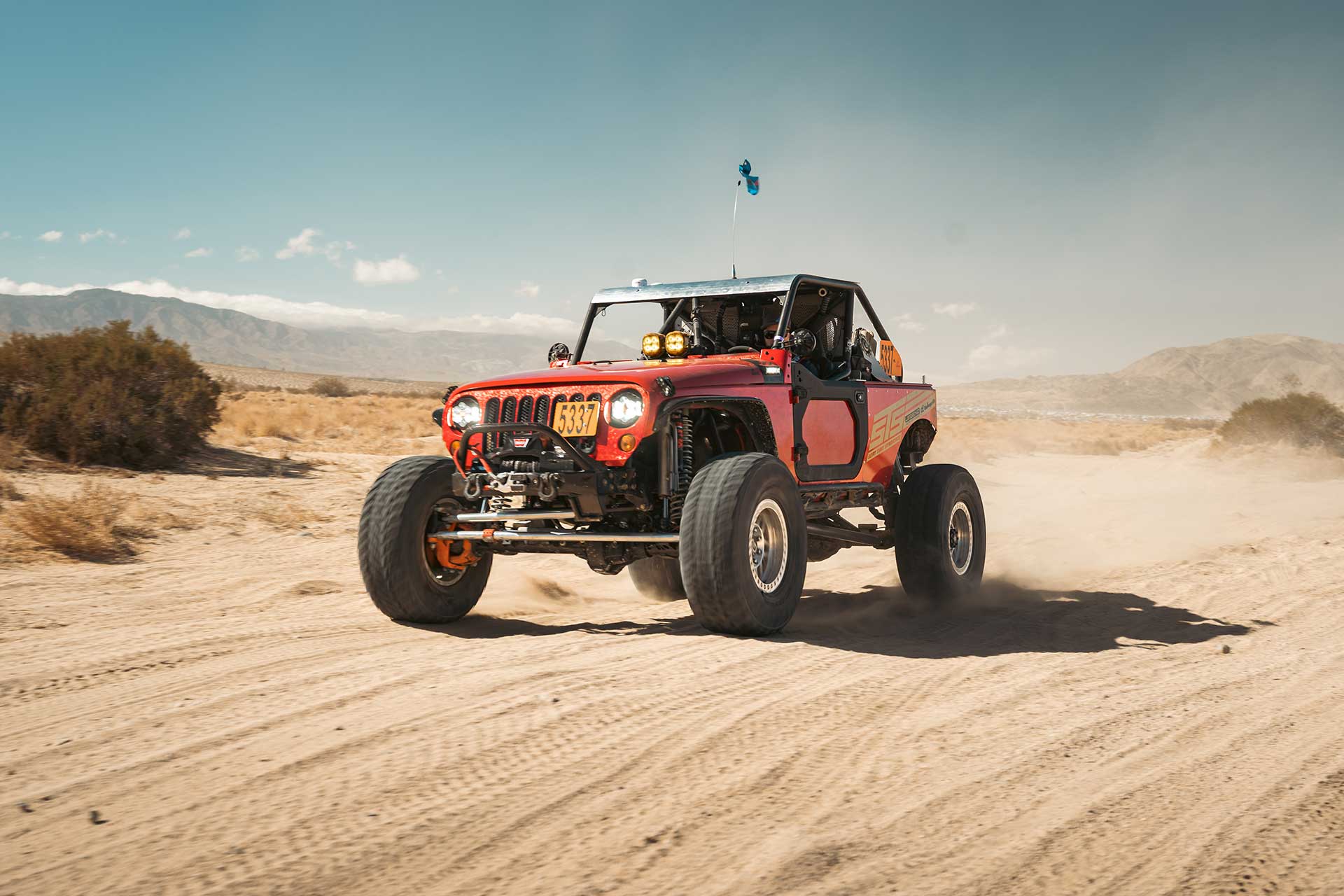 red off-road racing jeep wrangler jk with rbp repulsor mt3 tires driving through desert terrain with mountains in background during king of the hammers race